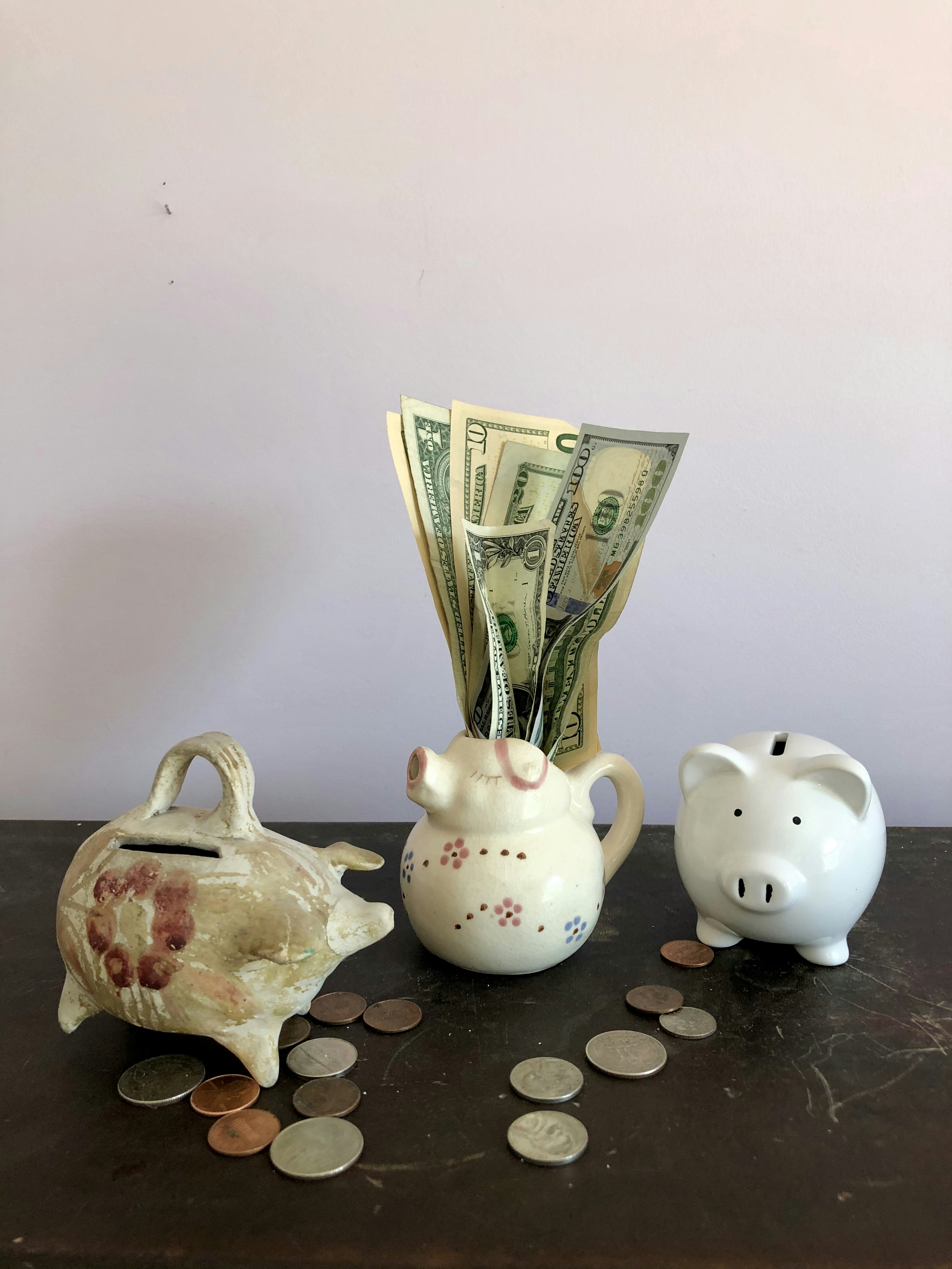 Two ceramic piggy banks and a small ceramic pig pitcher in a row on a wooden cabinet against a light lavender grey wall. US coins are scattered on the cabinet. A cluster of US paper currency in a variety of denominations sprout from the pig pitcher.